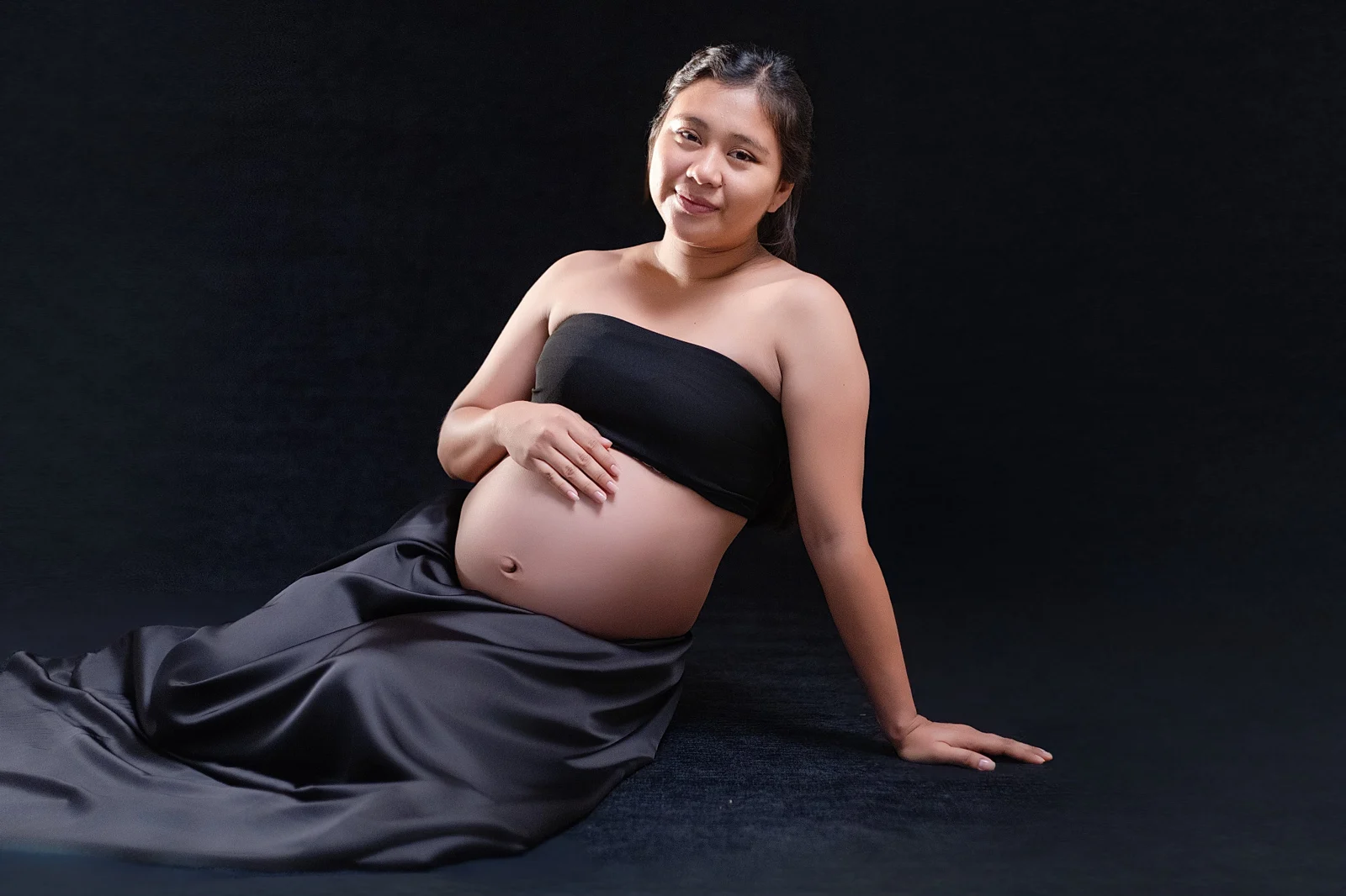 Mamá embarazada sentada con falda de satín negro, retrato de estudio